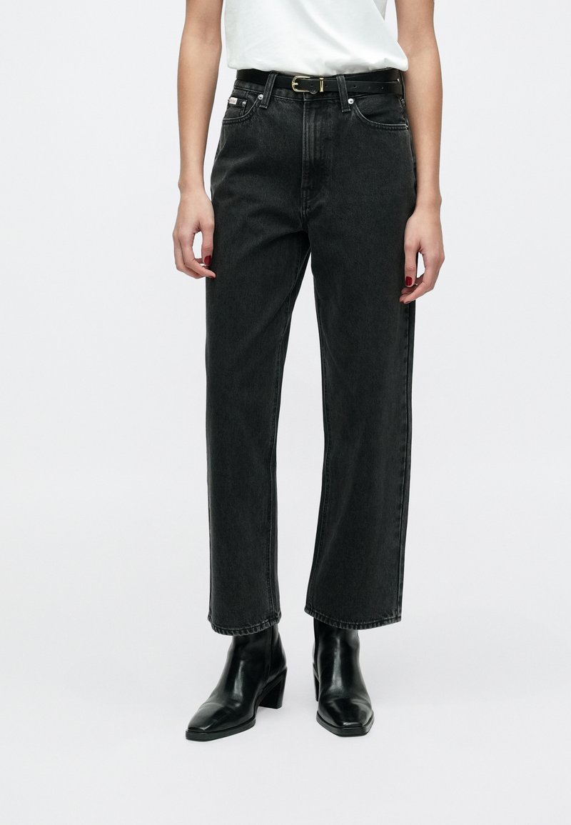 Jeansi negri din denim, talie înaltă, croială dreaptă și textură subtilă, asortați cu o curea neagră și cizme negre cu toc la gleznă.