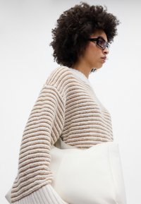 Raidallinen neulepaita beige ja valkoinen, suurilla hihoilla, yhdistettynä kermanväriseen laukkuun. Tekstuuri tuo visuaalista mielenkiintoa.