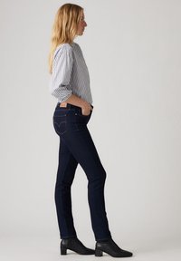 Jeans skinny azul oscuro con cintura alta, bolsillos planos y un sutil diseño de costura. Combinados con una camisa de botones rayada clara y botas de tobillo negras.