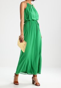 Grön, ärmlös maxiklänning med en samlad halsringning och en insnörd midja. Slät tyg och ankel-lång design. Parat med en gyllene kuvertväska och bruna klacksandaler.