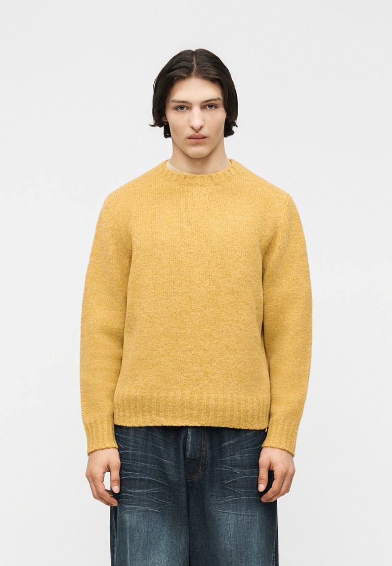 Jeune personne aux cheveux bruns mi-longs portant un pull en maille jaune moutarde et un jean large bleu foncé sur fond blanc.