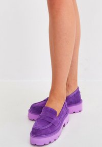 Mocassini in pelle scamosciata viola con suola spessa di lavanda, caratterizzati da dettagli cuciti e design slip-on. La texture è morbida e liscia.