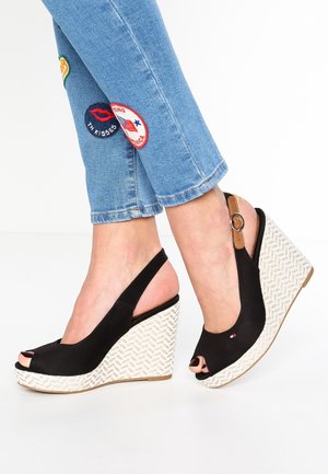 Fødder iført sorte peep-toe kilehælssandaler med flettede hvide hæle, kombineret med blå jeans med farverige lapper på underbenene.