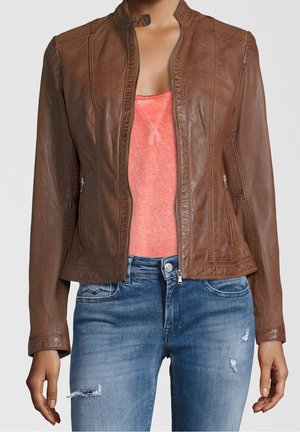 Frau trägt eine braune Lederjacke über einem korallenfarbenen Tanktop und zerrissene blaue Jeans, steht vor einem schlichten hellen Hintergrund.