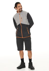 Giacca in pile grigia e nera con zip arancione e dettagli arancioni, abbinata a pantaloni corti scuri e scarpe da trekking nere. Texture liscia, vestibilità atletica.