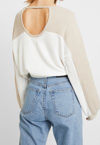 Pull en tricot à côtes beige et blanc avec une découpe dans le dos, doté de manches longues, d'une coupe loose et associé à un jean bleu taille haute.