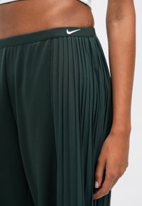 Calças de atletismo plissadas Nike em verde escuro com uma cinta elástica lisa, apresentando um pequeno logótipo branco. Tecido texturizado com pregas verticais.