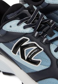 Baskets de sport bleu marine et clair présentant un dessus en tissu texturé, des accents brillants, des lacets et un logo proéminent sur le côté.