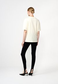 Moteris stovi atsukusi nugarą, vilkėdama laisvą baltą marškinėlį, juodas prigludusias legingas su užkulniais ir juodas aukštakulnius atviras batus.