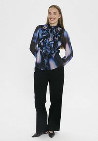 Caura zila un melna blūze ar rumbu priekšpusi, apvienota ar melnām platām biksēm un melniem norādītā purngalā apaviem.