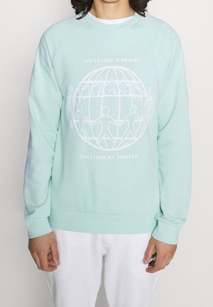 Personne portant un sweat-shirt bleu clair avec un globe blanc et des personnages en bâton se tenant la main, le texte indique "The future is bright, together we succeed."