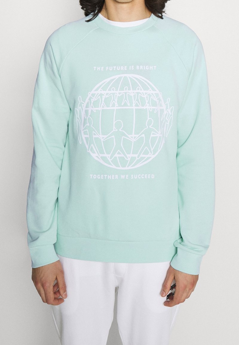 Personne portant un sweat-shirt bleu clair avec un globe blanc et des personnages en bâton se tenant la main, le texte indique "The future is bright, together we succeed."