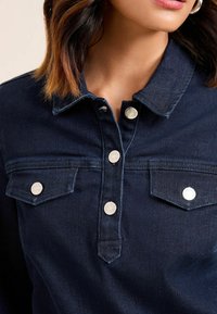 Gros plan sur une personne portant une veste en denim bleu foncé avec des boutons-pression en argent et des poches à rabat sur la poitrine.