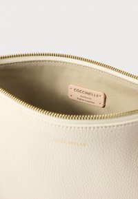 Krémfehér bőr tasak texturált felülettel, arany cipzárral, bézs béléssel és "Coccinelle - Eredeti olasz bőr" feliratú címkével.