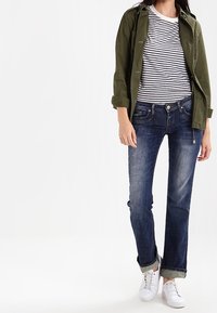 Kurtka w oliwkowej zieleni na granatowo-białej, paskowanej koszulce, do tego jasne, proste dżinsy i białe trampki. Dżinsy podwinięte, widoczne kieszenie.