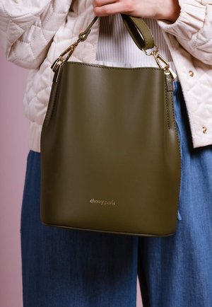 Personne tenant un sac seau en cuir vert olive avec des ferrures dorées, portant une veste matelassée crème et un pantalon bleu.