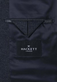 Tmavá vzorovaná látka s trojúhelníkovým klopou a černou obdélníkovou štítkem značky Hackett London přišitým pod ní.