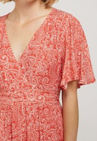 Robe à motif floral rouge avec de courtes manches ondulées, un décolleté en V profond et une taille nouée. Le tissu a une texture douce avec des détails paisley complexes.