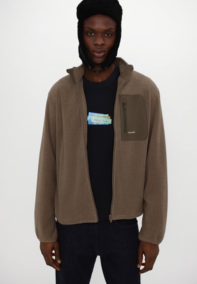 Braune Fleecejacke mit einer Reißverschlusstasche auf der linken Brust, kombiniert mit einem marineblauen T-Shirt mit einem bunten Grafikdesign.