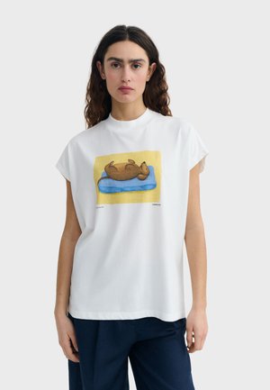 Femme aux longs cheveux foncés portant un t-shirt blanc avec une illustration d'un chien brun allongé sur un lit bleu avec un fond jaune.