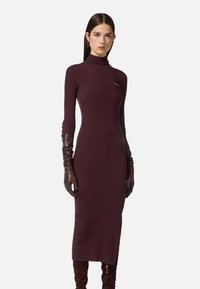 Abito a coste color bordeaux con collo alto, spacco laterale e una piccola tasca sul petto. Abbinato a guanti lunghi in pelle marrone liscia.