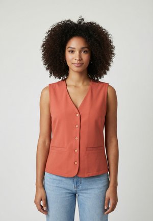 Donna con capelli ricci che indossa un gilet senza maniche color ruggine con bottoni e jeans azzurri, in piedi contro uno sfondo semplice.