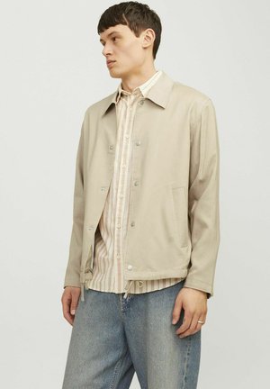 Beige canvas jack met een kraag, drukknopen, zijzakken en een verstelbare onderkant. Draagt over een gestreepte blouse en dennenbroek.