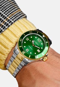 Grønn dykkerklokke med bezel i gullakenter, sølv- og gullmetallarmbånd, hvite timeindekser og et datovindu ved 3-tiden.