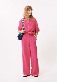 Helderroze, gestructureerde katoenen tweedelige outfit met een geknoopte blouse, losse broek en een kleine blauwe geweven tas met een gevlochten handvat.
