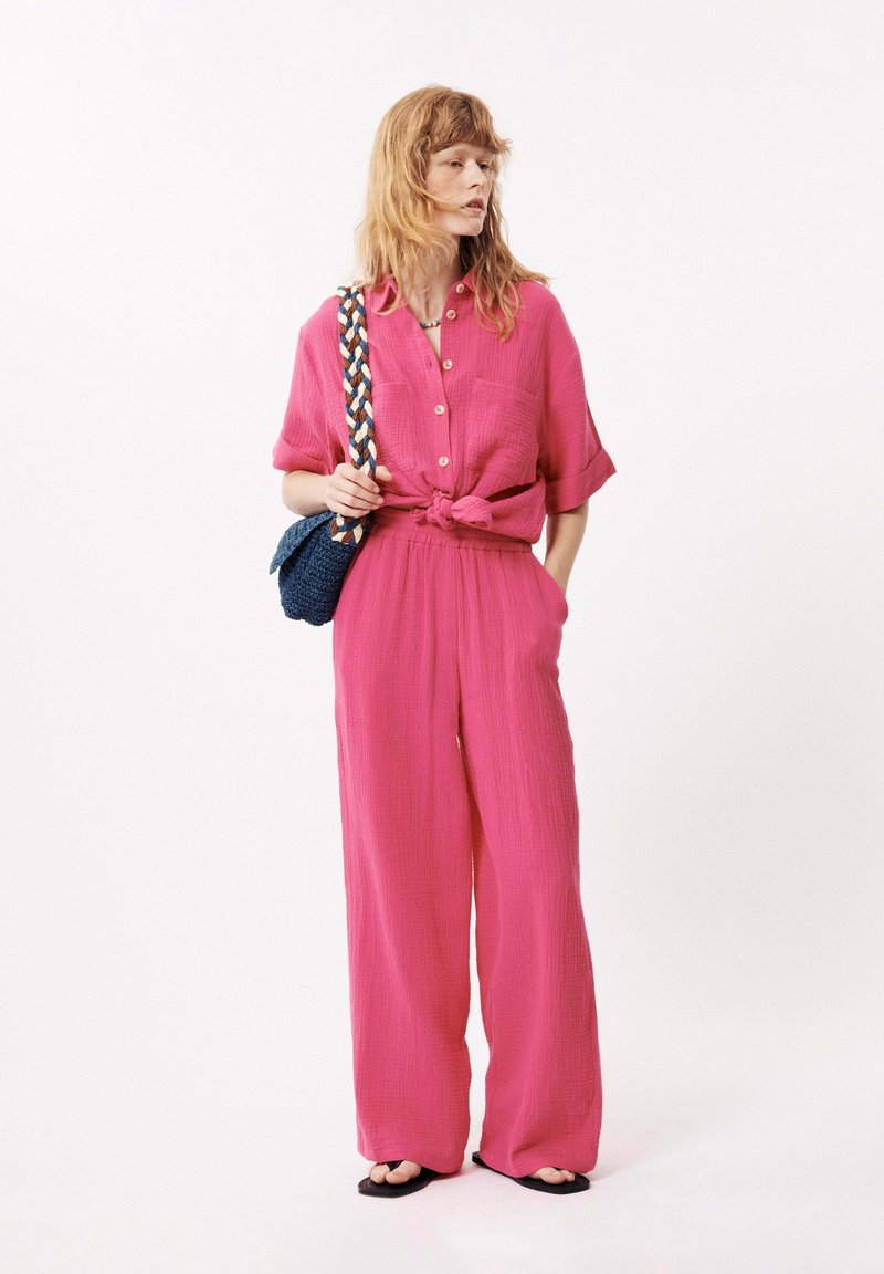 Helderroze, gestructureerde katoenen tweedelige outfit met een geknoopte blouse, losse broek en een kleine blauwe geweven tas met een gevlochten handvat.