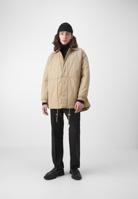 Beige pufferjack met een rits en drukknopen aan de voorkant, elastische manchetten en een trekkoord onderaan. Draagt over een zwarte trui, gecombineerd met zwarte broeken en schoenen.