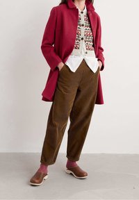 Abrigo rojo con textura, chaleco tejido con estampado, camisa blanca, pantalones de pana marrones, calcetines rosas y zuecos marrones forman un conjunto casual y con capas.