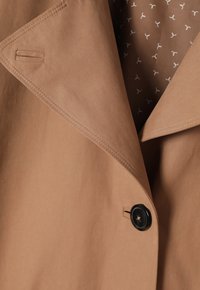 Trench-coat beige avec un grand col, un bouton noir unique et des détails de couture. Doublé d'un motif subtil sur fond marron.