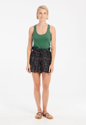 Grünes Tanktop mit Spitzenschultern, kombiniert mit schwarzen Shorts mit floralem Muster und elastischem Bund. Das Model trägt außerdem flache Sandalen.