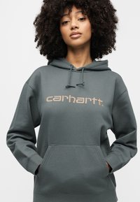 Σκούρα πράσινη μπλούζα με κουκούλα από μαλακό ύφασμα, με τσέπη καγκουρό και κουκούλα με κορδόνι. Διαθέτει το λογότυπο «carhartt» σε πιο ανοιχτό χρώμα.