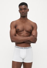Weisse Baumwoll-Boxershorts mit einem schwarzen Bund, der mit wiederholtem Text bedruckt ist. Das Modell posiert mit verschränkten Armen und präsentiert die Passform der Shorts.