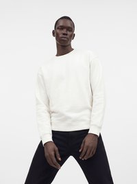 Homme portant un sweat-shirt blanc uni et un pantalon noir, debout les jambes écartées sur fond blanc.