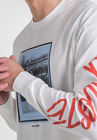 Camicia bianca a maniche lunghe caratterizzata da una stampa fotografica con immagini storiche e testo rosso audace sulla manica sinistra. Tessuto morbido con superficie liscia.