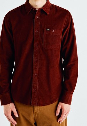 Homme portant une chemise en velours côtelé rouge foncé à manches longues avec une poche boutonnée et un pantalon marron, les mains détendues le long du corps.