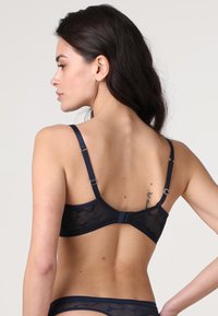 Reggiseno in pizzo blu marina con spalline regolabili, fascia liscia e motivo floreale su materiale trasparente. Chiusura posteriore con gancio e occhiello.