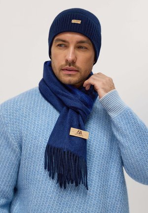 Berretto in maglia navy e sciarpa coordinata con estremità frangiate. La sciarpa presenta un'etichetta in tessuto. Il maglione è di un blu chiaro con un design texture.