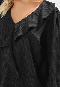 Blusa nera texturizzata con scollatura e maniche a volant. Il tessuto presenta un design plissettato con una leggera lucentezza.