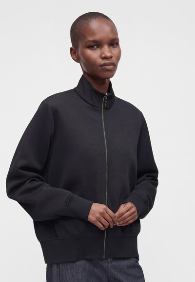 Personne aux cheveux courts portant une veste noire zippée et un pantalon bleu, debout avec les mains jointes devant un fond gris uni.
