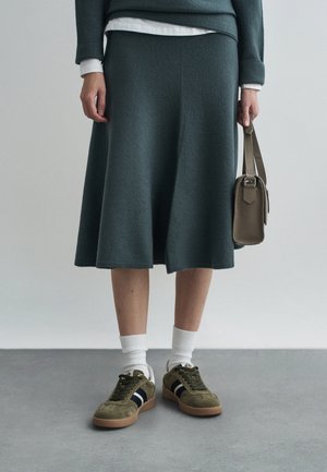 Gonna verde a maglia con una forma svasata, abbinata a sneaker in suede verde oliva con strisce nere e calzini bianchi. Piccola borsa in mano.