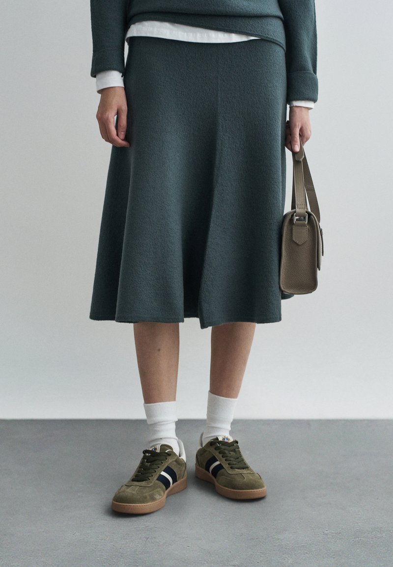 Πλεκτή πράσινη φούστα σε φαρδιά γραμμή, συνδυασμένη με ελαιοπράσινα σουέντ αθλητικά παπούτσια με μαύρες ρίγες και άσπρες κάλτσες. Μικρή τσάντα στο χέρι.