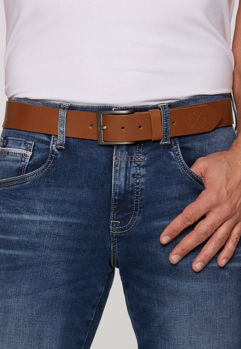 Brauner Ledergürtel mit einer rechteckigen Metallschnalle, getragen mit dunklen Denim-Jeans, hat eine flache Textur und mehrere Einstelllöcher.