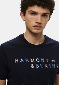 Maglietta in cotone blu navy con logo riflettente "HARMONT & BLAINE" e una piccola grafica di un cane bianco. Collo classico a girocollo e maniche corte.