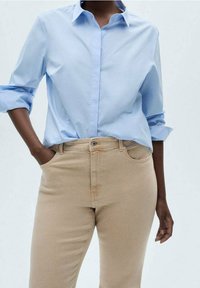 Personne portant une chemise bleu clair à manches longues retroussées et un pantalon beige, une main dans la poche, sur un fond uni.