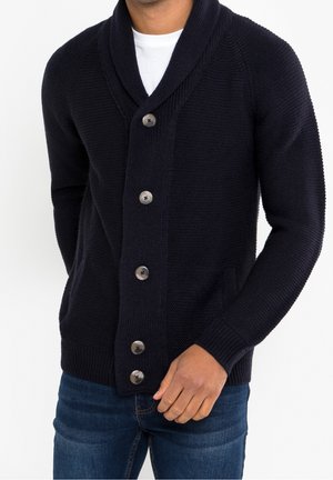 Homme portant un cardigan en tricot noir texturé avec de gros boutons par-dessus une chemise blanche et un jean bleu, debout avec une main près de la taille.
