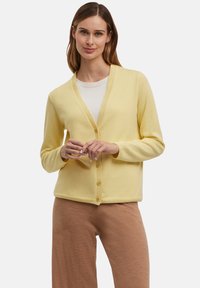 Geel vest met een V-hals, vijf knopen en lange mouwen. De stof lijkt zacht en lichtgewicht, gecombineerd met beige broeken.